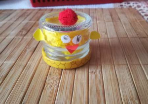 Kurczaczek wykonany przez dziecko z małego słoiczka z doklejonymi z żółtego papieru skrzydełkami oraz oczkami i z czerwoną na głowie kuleczką